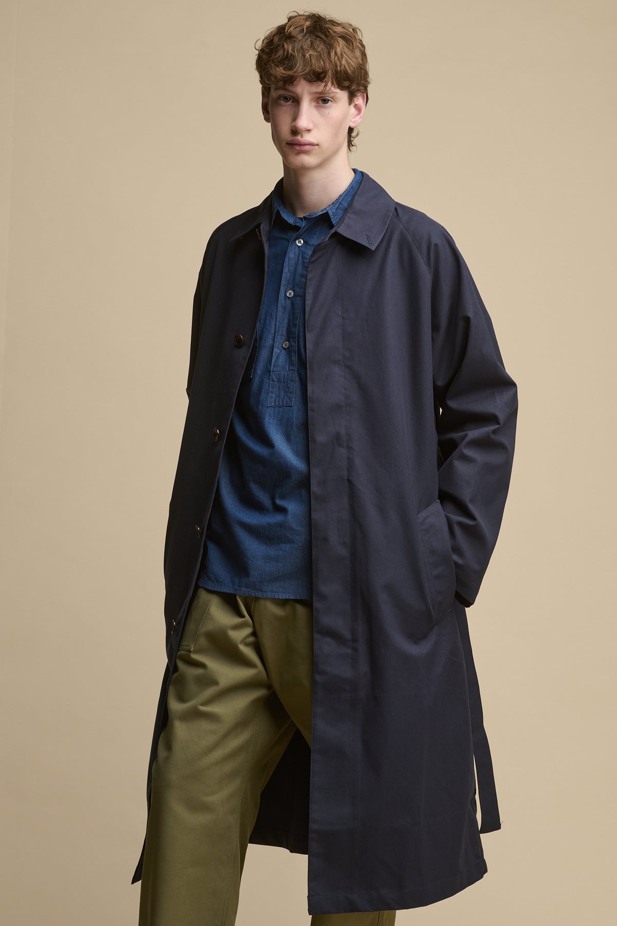 Full body image of male wearing Oli Half Placket Lightweight Denim Shirt in indigo paired with cameraman pants in olive and men's Frank raglan belted raincoat in indigo