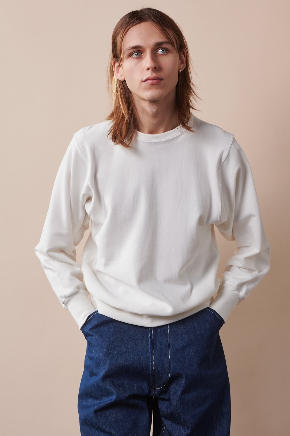 
            Thigh up image of male with shoulder length hair wearing 80s sweatshirt in bone, with hands in pockets of blue jeans