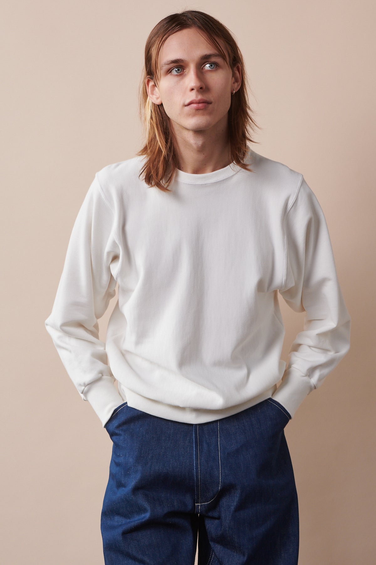 Thigh up image of male with shoulder length hair wearing 80s sweatshirt in bone, with hands in pockets of blue jeans