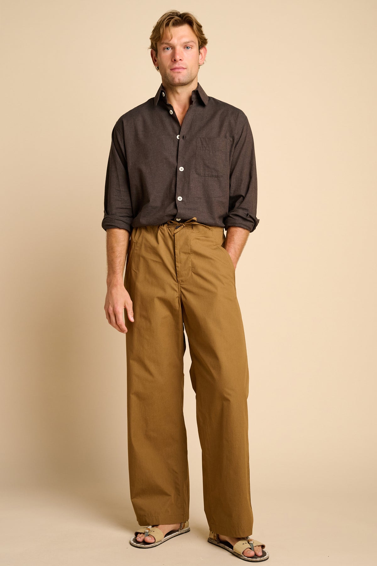 Man wearing tan trousers with brown shirt against beige background.