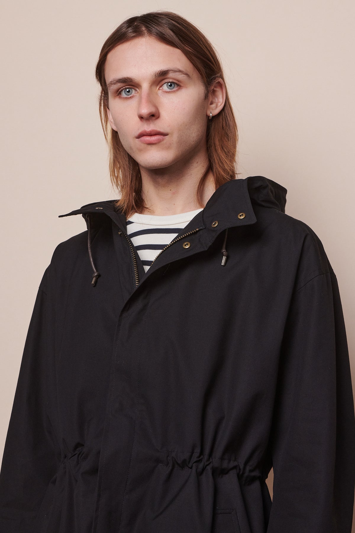 Portrait of male wearing parka in black with waist drawstring tightened to synch waist. Adjustable hood drawstring visible.