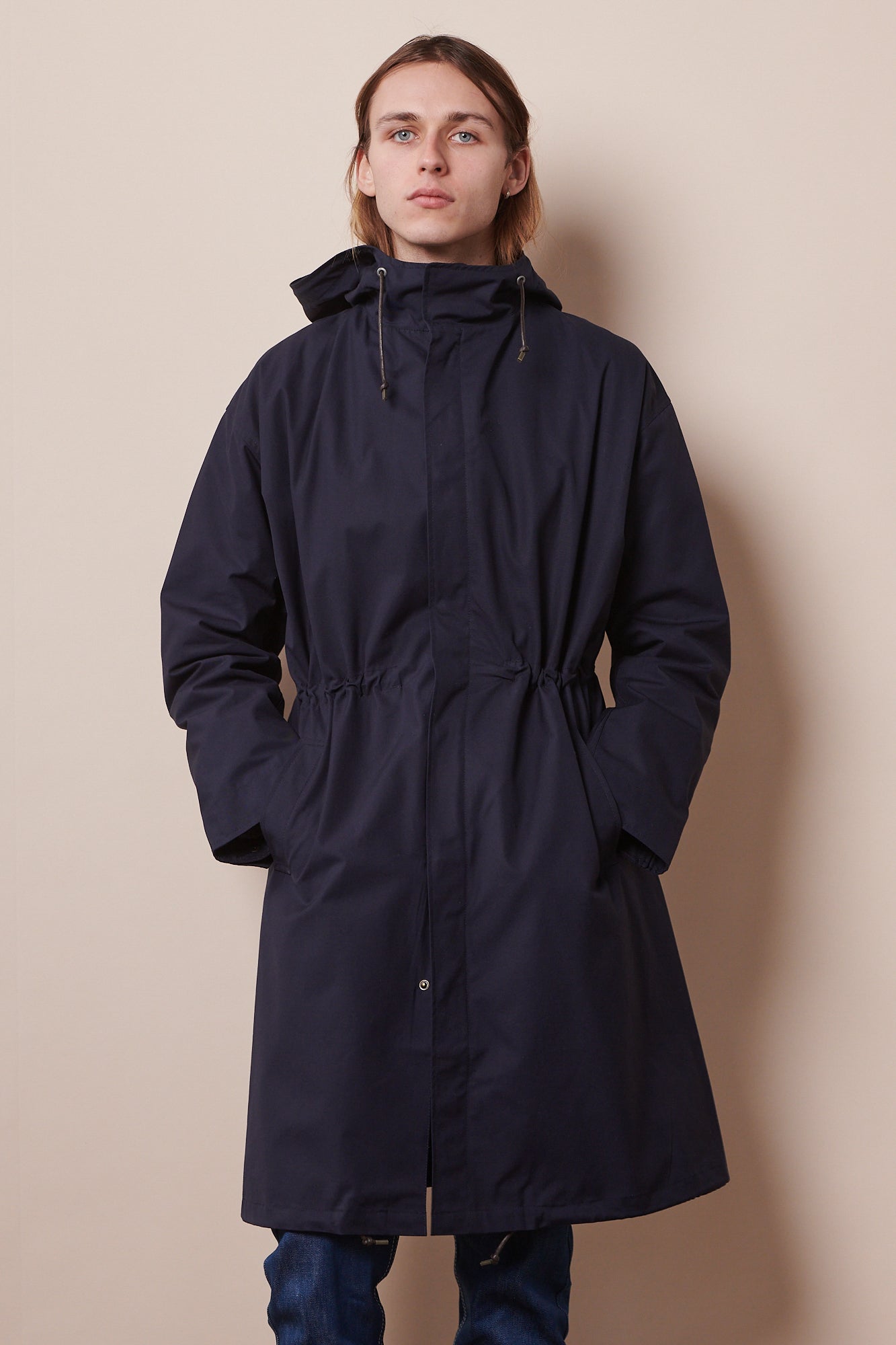 Knee up image of the front of male wearing zipped parka in navy with hands in front pockets. 