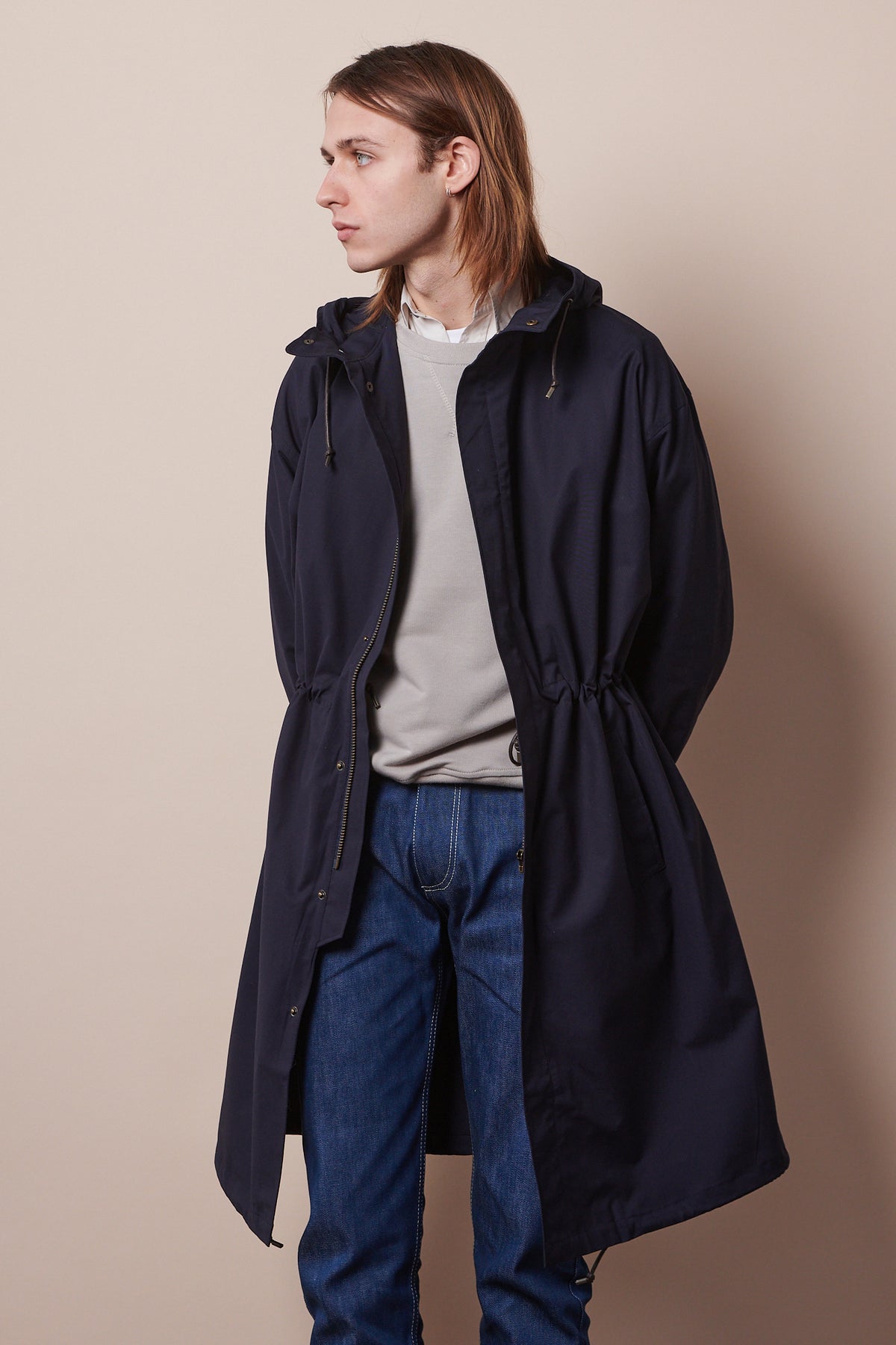 Knee up image of male looking to the side wearing parka in navy unzipped, layered over stone raglan sweatshirt and blue jeans