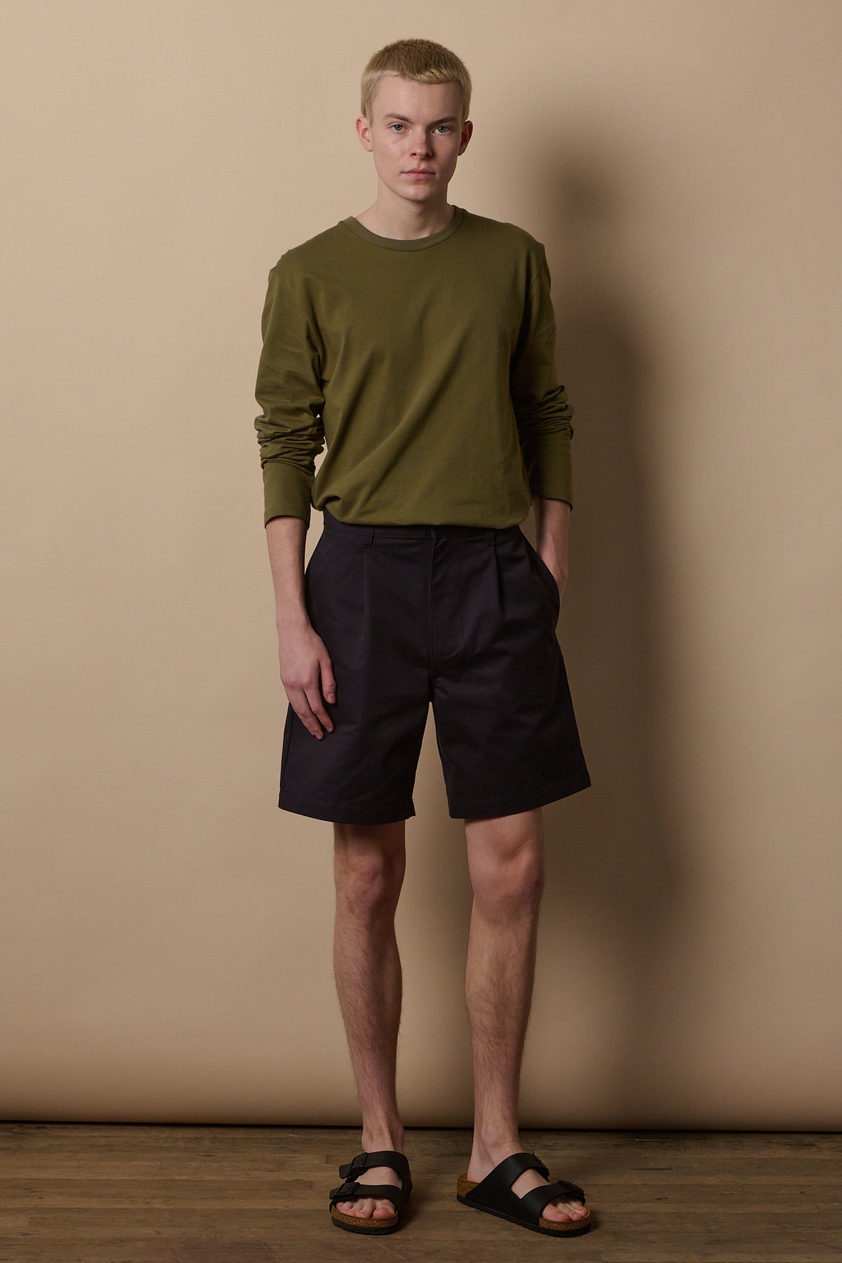 
            White male wearing pleated shorts in navy paired with long sleeve t shirt in olive tucked in.