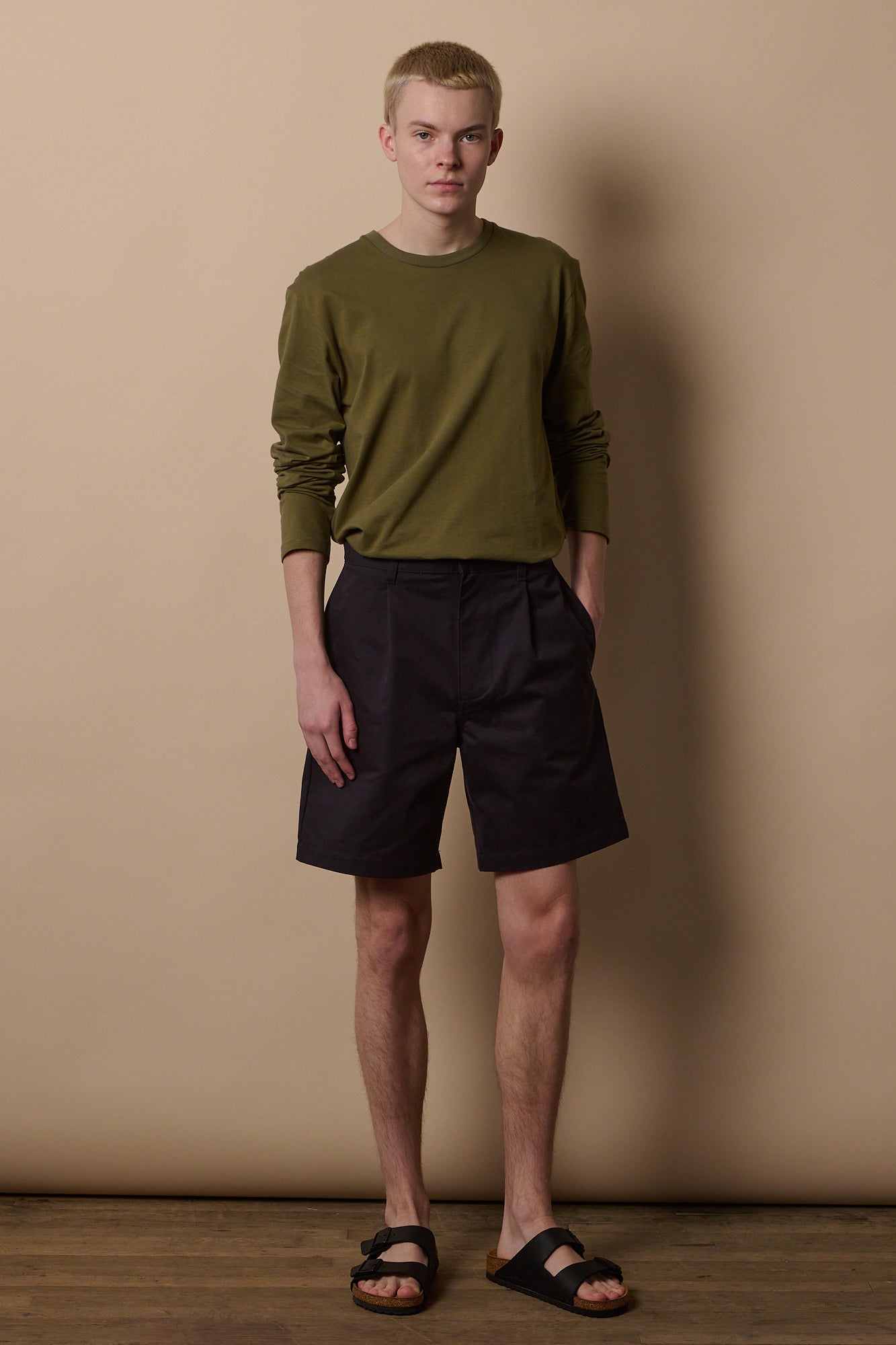 White male wearing pleated shorts in navy paired with long sleeve t shirt in olive tucked in.