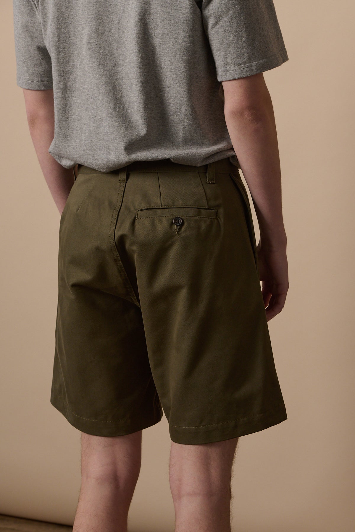Back of male wearing pleated shorts in olive showing back pocket with button fastening, and belt loops on the waistband