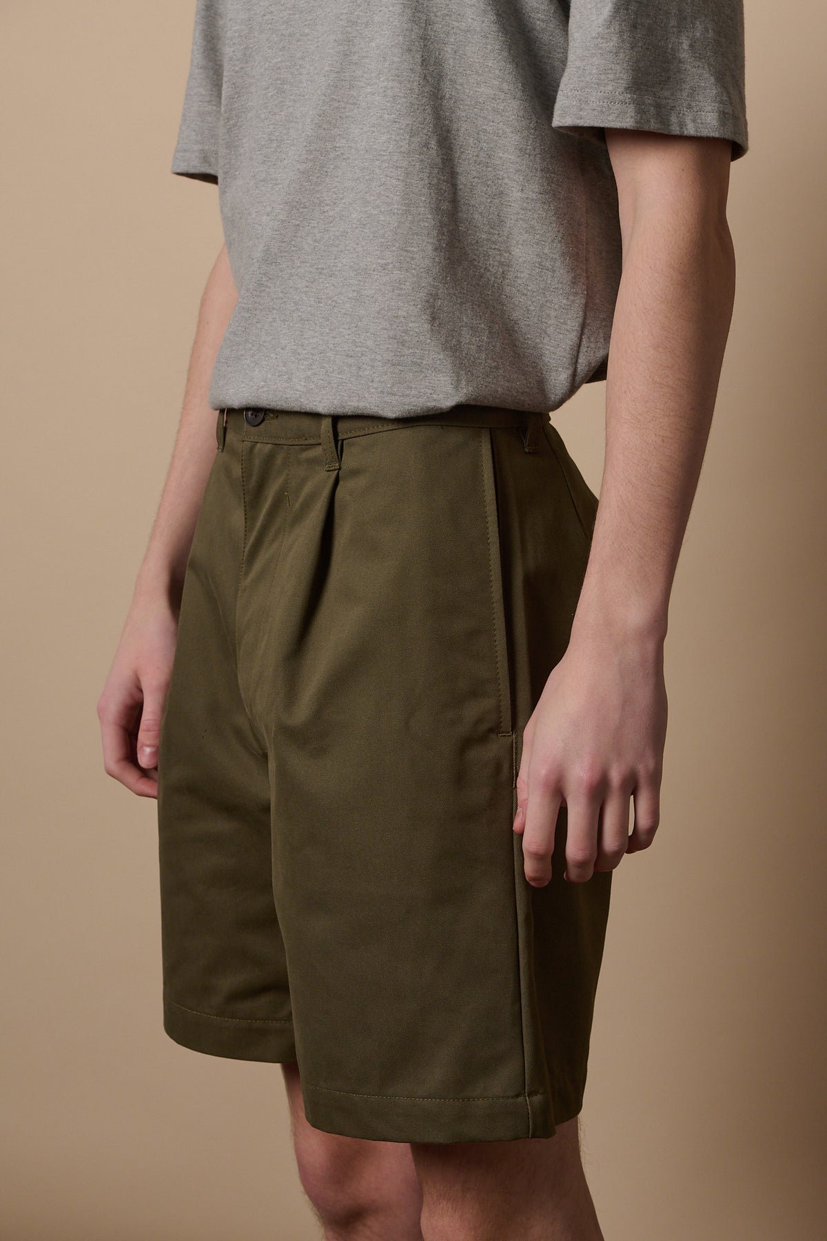 Image of white male wearing pleated shorts in olive paired with grey short sleeve t shirt tucked in