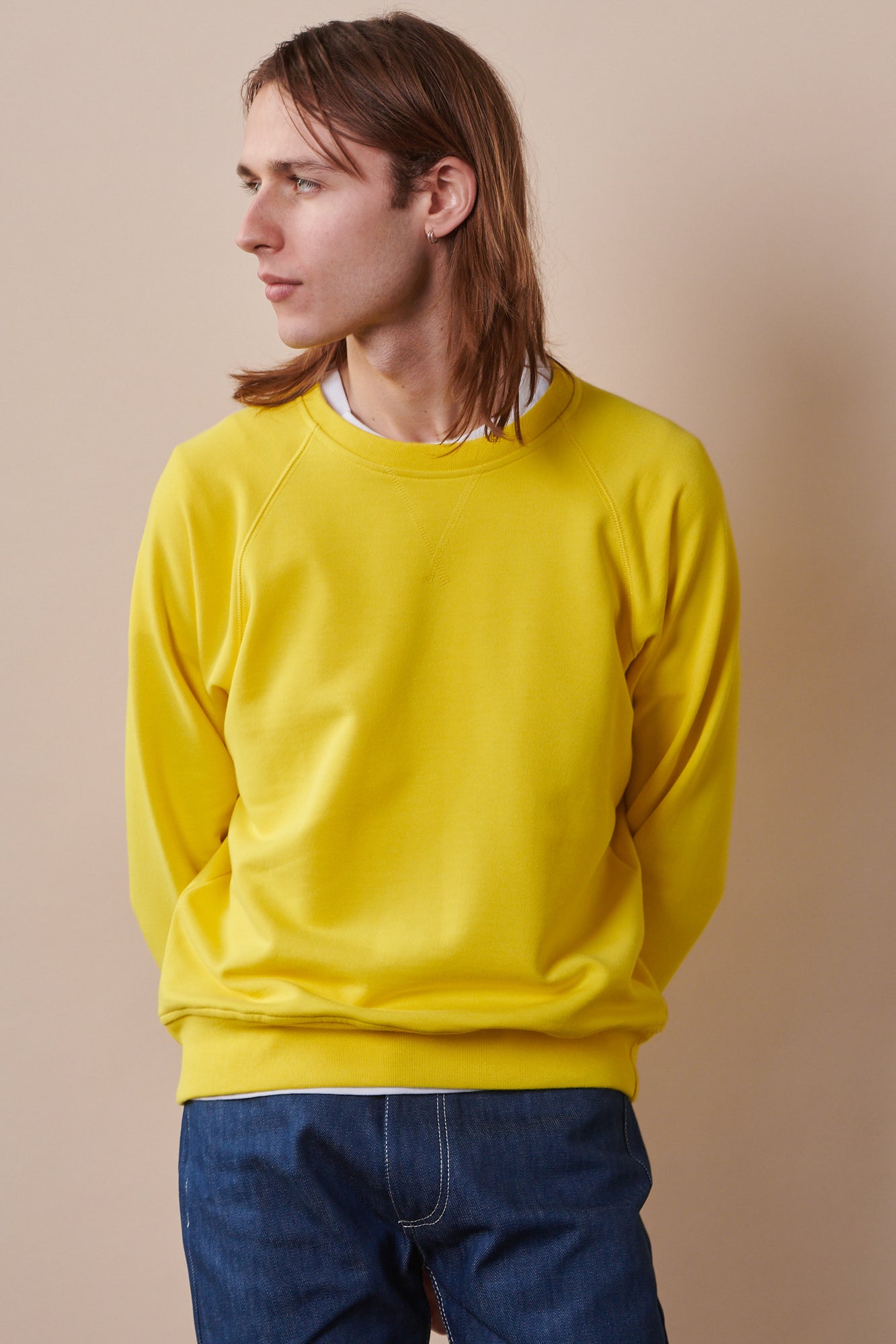 Male with shoulder length brown hair wearing raglan sweatshirt in canary yellow paired with blue jeans.