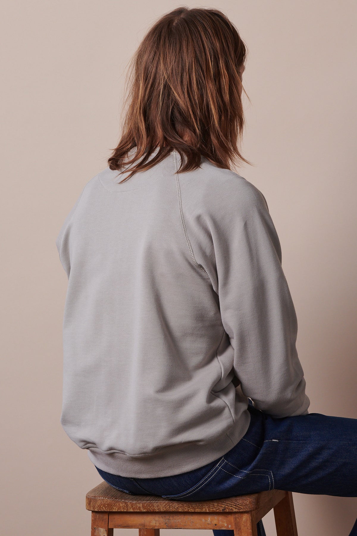 Image of the back of male with shoulder length brown hair sat on stool wearing raglan sweatshirt in stone