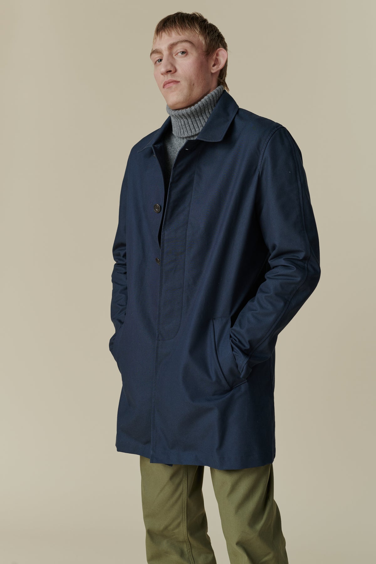 Side angle of male wearing raincoat in navy hands in two front pockets, mid thigh length.