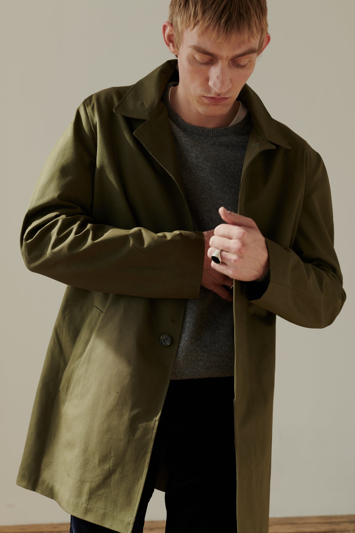 
            Thigh up image of male wearing men&#39;s raincoat in olive. 2 side welt pockets. Concealed button placket. Half lined