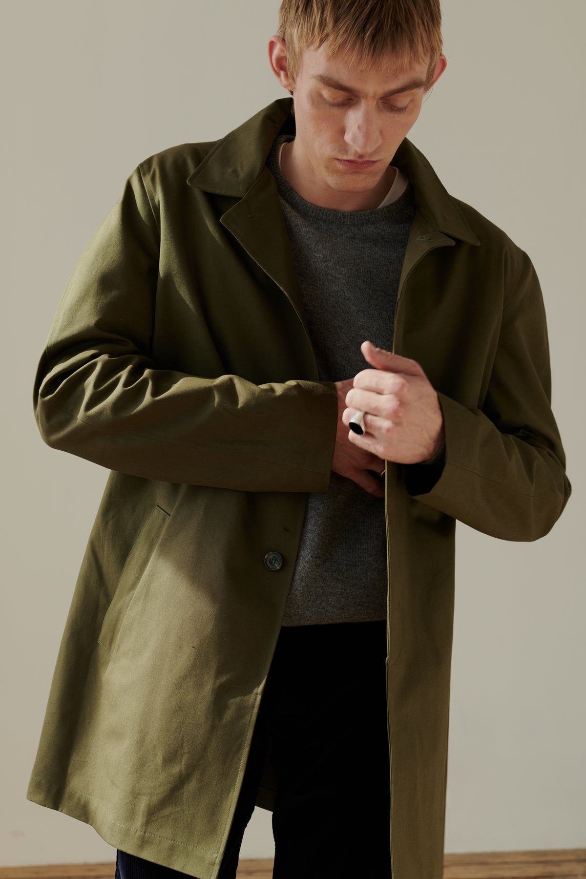 Thigh up image of male wearing men's raincoat in olive. 2 side welt pockets. Concealed button placket. Half lined