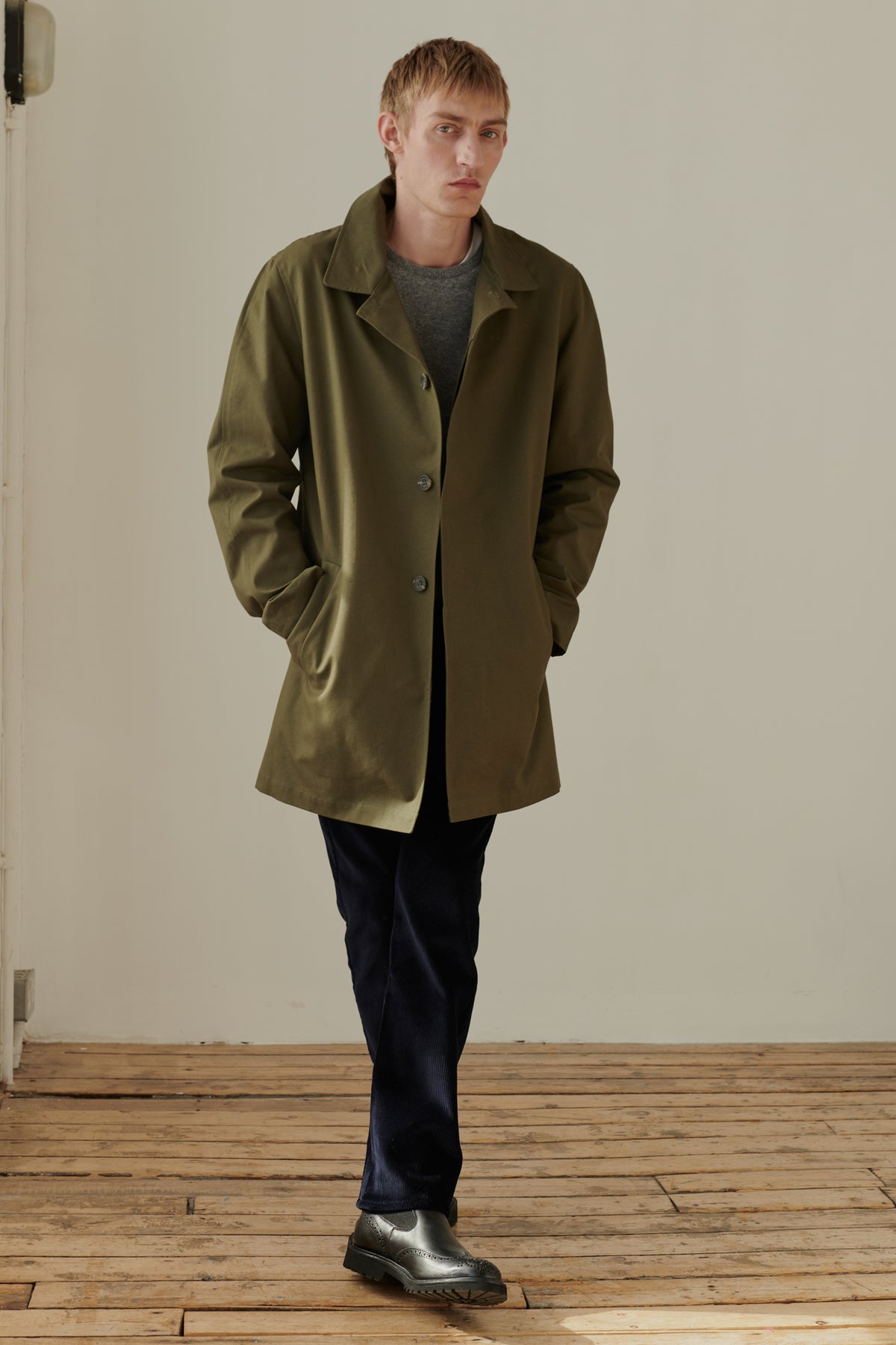 Full body image of male wearing men's raincoat in olive. 2 side welt pockets. Concealed button placket. Half lined