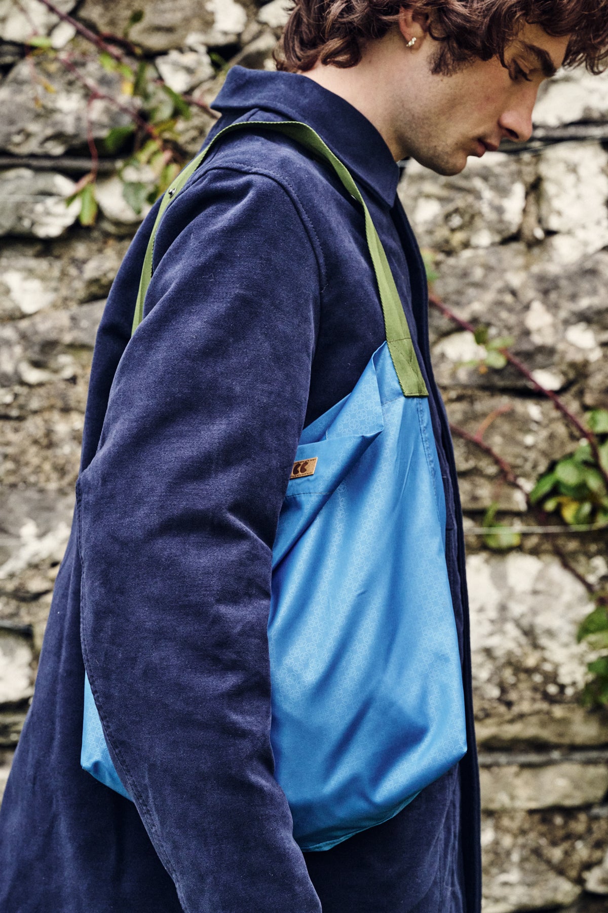 Person wearing a blue jacket with a blue bag over their shoulder against a stone wall background