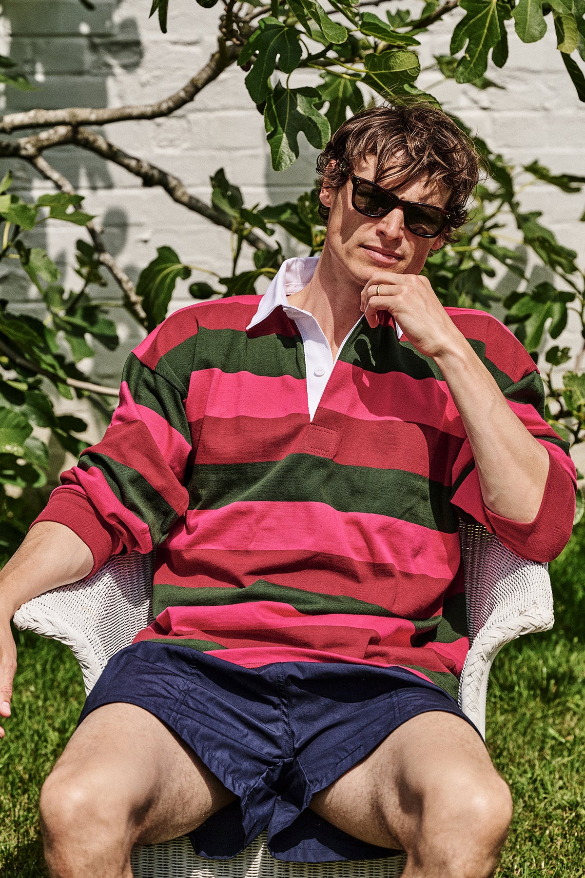 A man wearing pink and green striped rugby shirt and navy shorts sat in a chair outdoors against tree in the background.