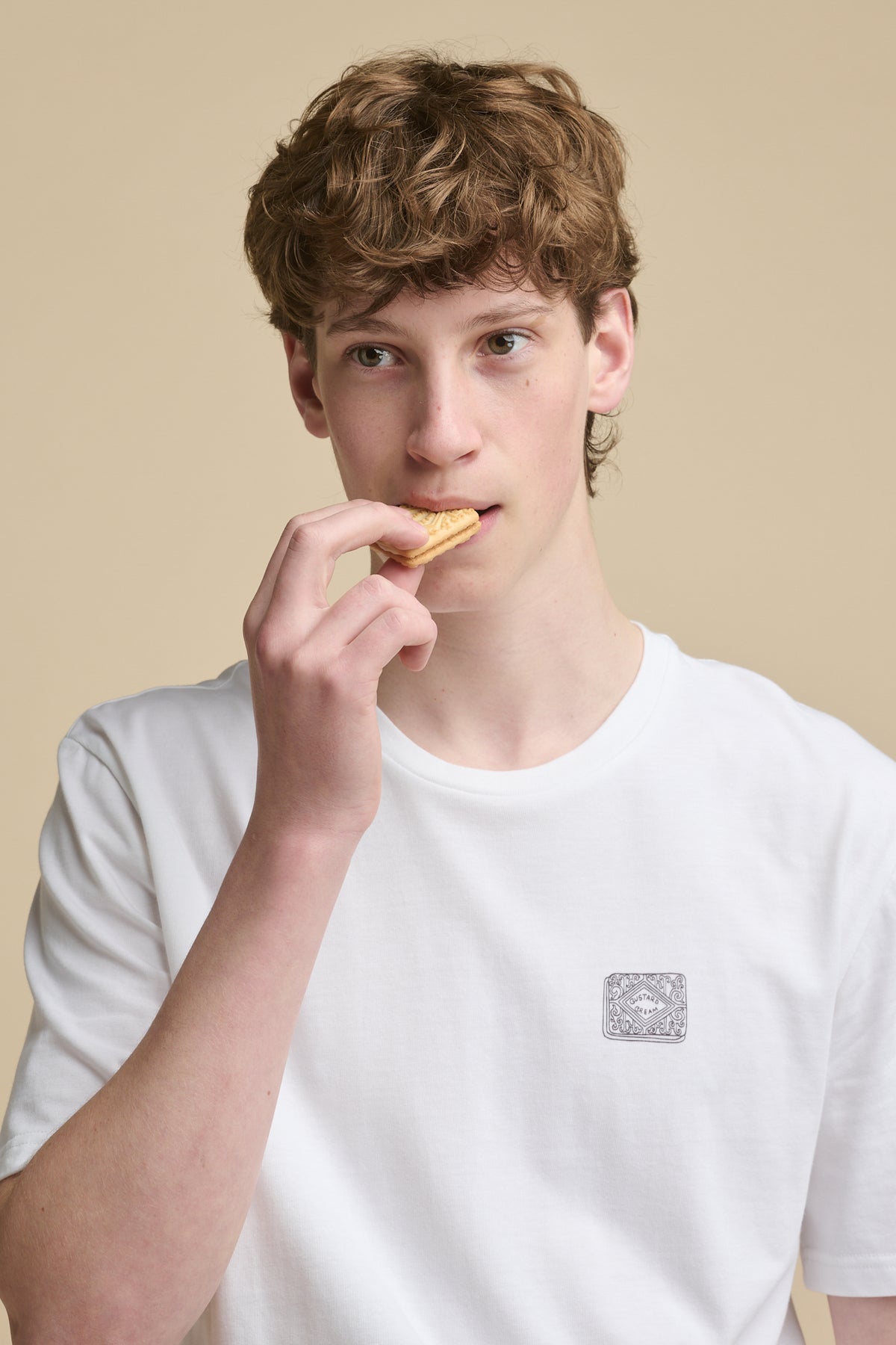 
            Portrait of male biting into custard cream biscuit whilst wearing short sleeve graphic t shirt in white with custard cream black outline print on