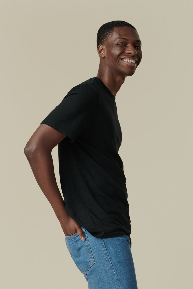 Thigh up image of smiley male from the side looking back towards the camera with his hand in his back pockets, wearing short sleeve t shirt plastic free in black with straight cut selvedge jeans in fade