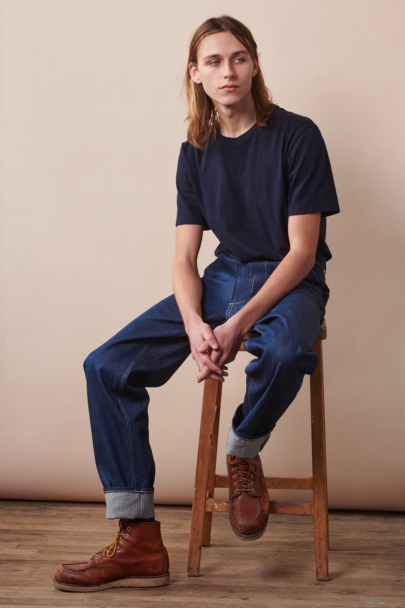 Full body image of male sat on stool with arms leaning on legs wearing short sleeve t shirt in navy paired with chore jeans in blue