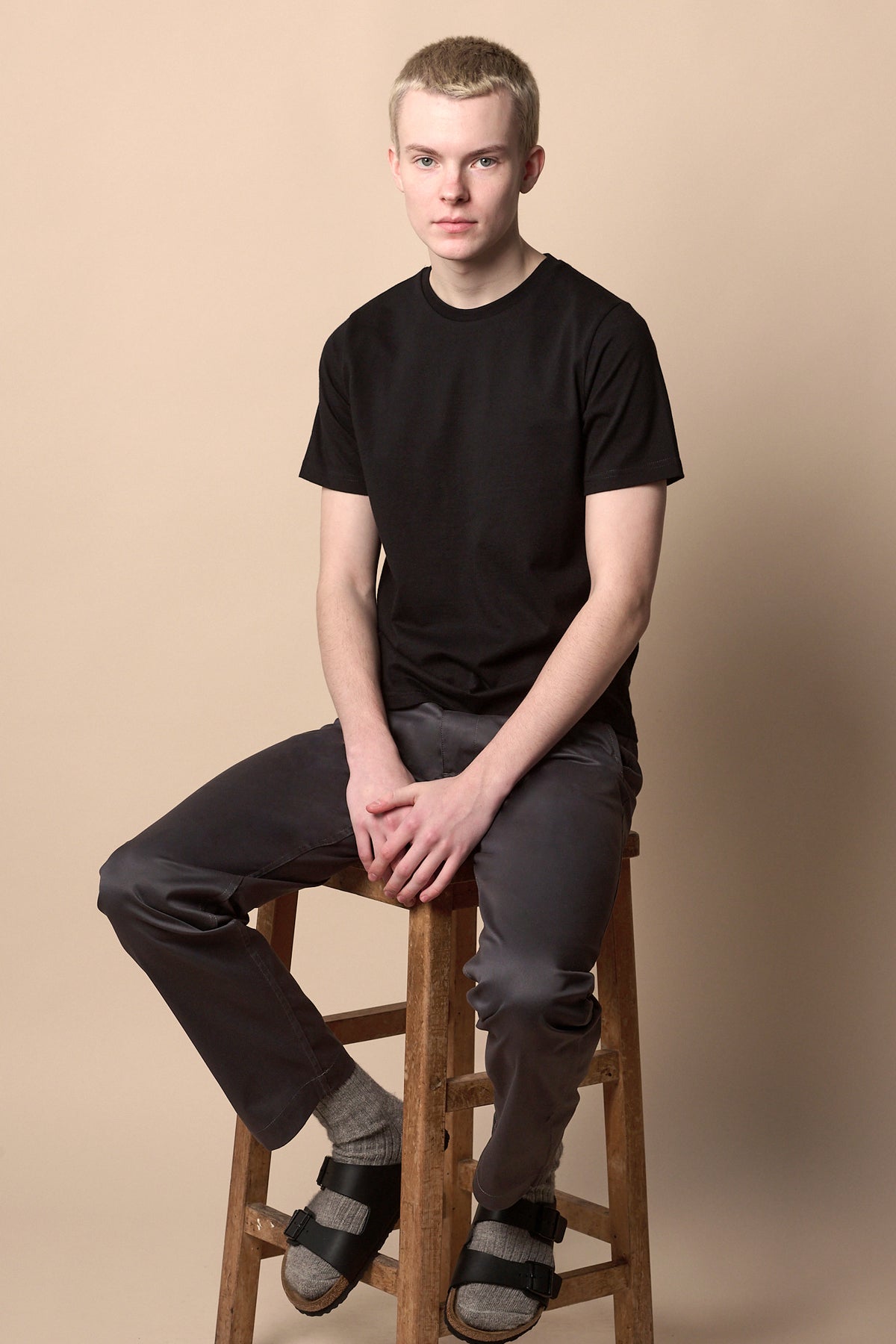 Male sat on stool wearing men's slim stretch chino in slate grey with black short sleeve crew neck t shirt and socks and Birkenstocks