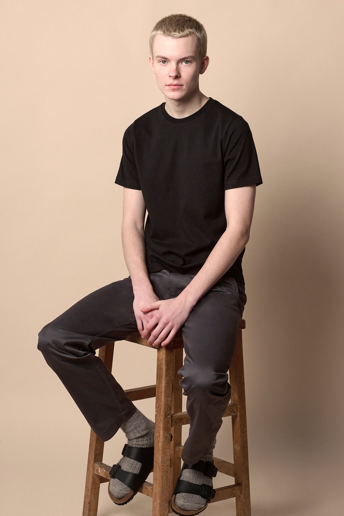 
            Male sat on stool wearing men&#39;s slim stretch chino in slate grey with black short sleeve crew neck t shirt and socks and Birkenstocks