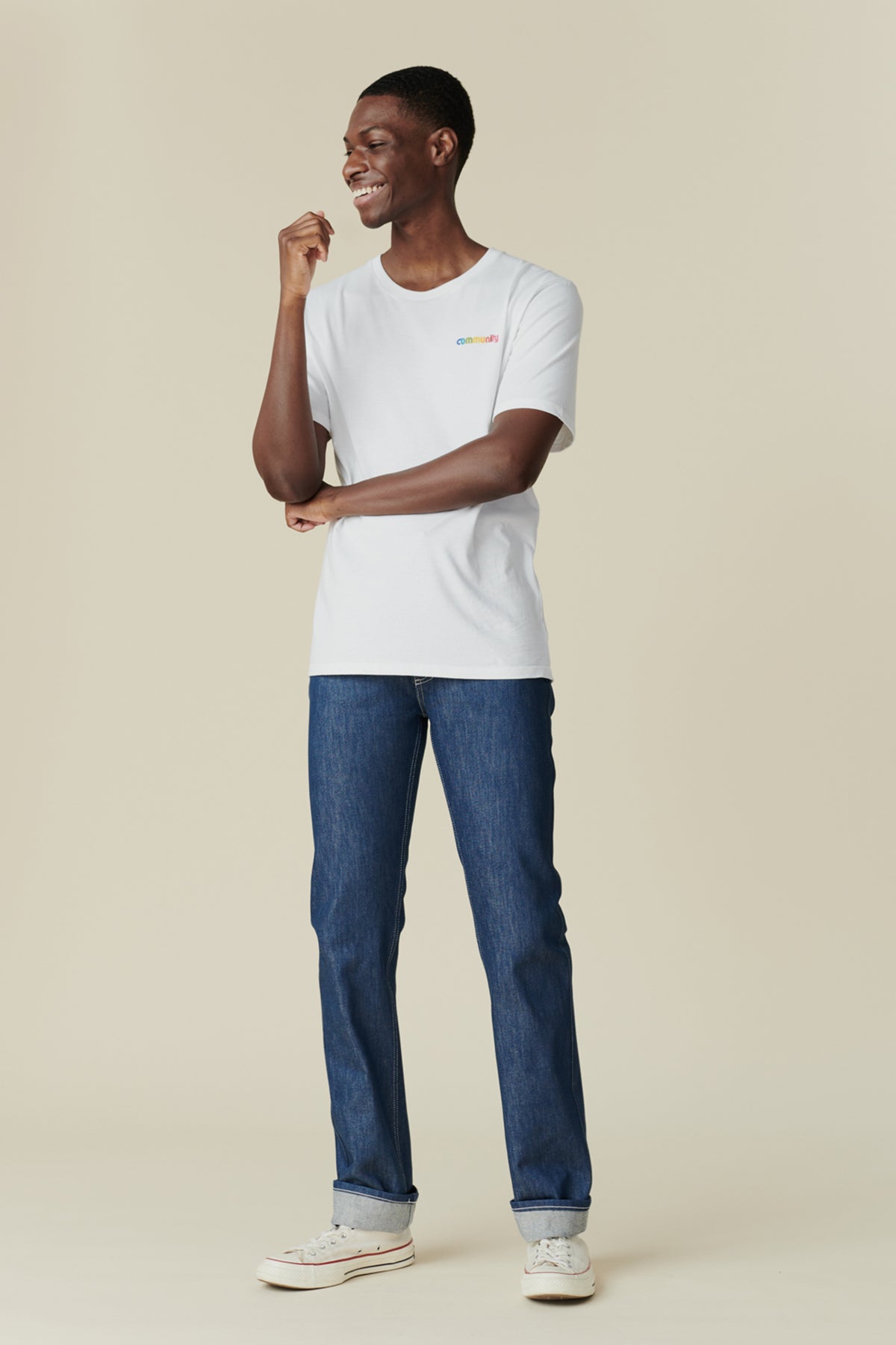A man standing against a beige background, wearing a white t-shirt and blue straight cut denim jeans with five pockets.