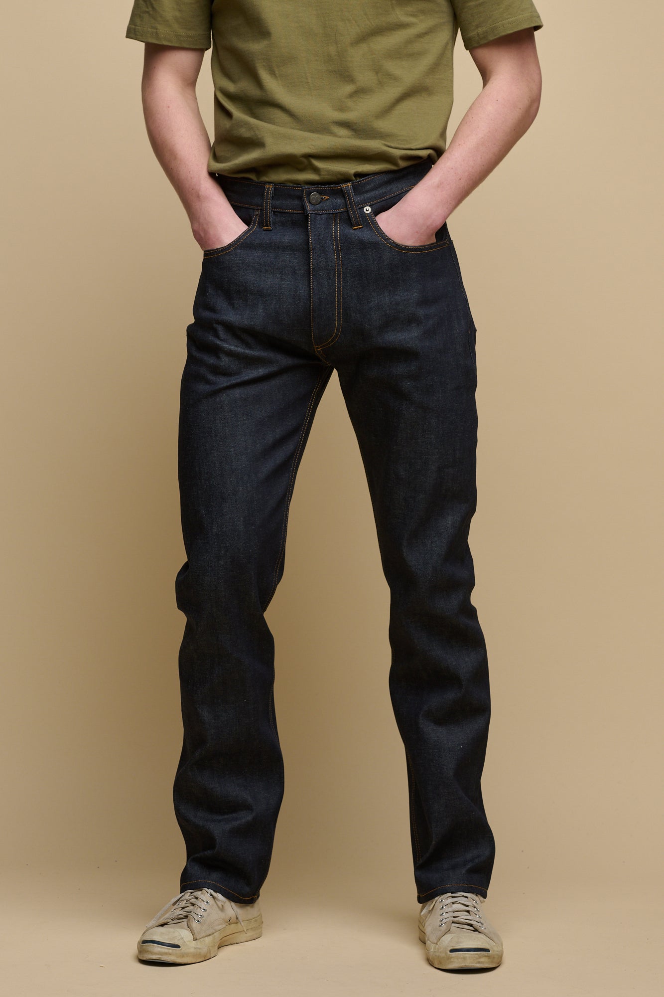 Chest down of male wearing men's straight cut selvedge jeans in raw denim with both his hands in the front pockets, paired with short sleeve t-shirt in olive tucked into the jean.