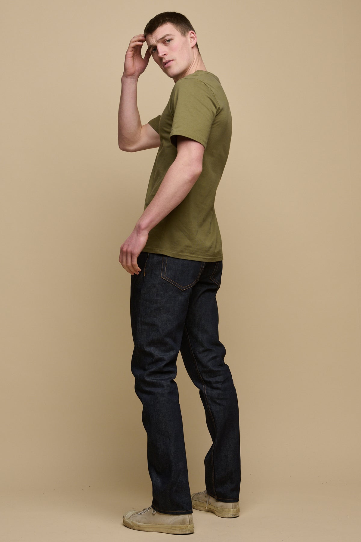 The back of male with his body facing away, looking back over his shoulder toward the camera wearing men's straight cut selvedge jeans in raw denim paired with short sleeve t-shirt in olive.