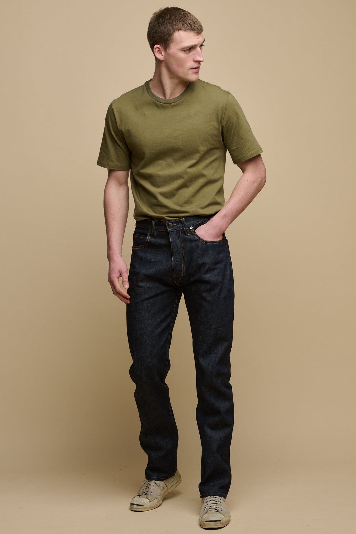Full body of the front of male looking to the side wearing straight cut selvedge jeans in raw denim with one of his hands in the front pocket paired with short sleeve t-shirt in olive tucked into the jeans with trainers.