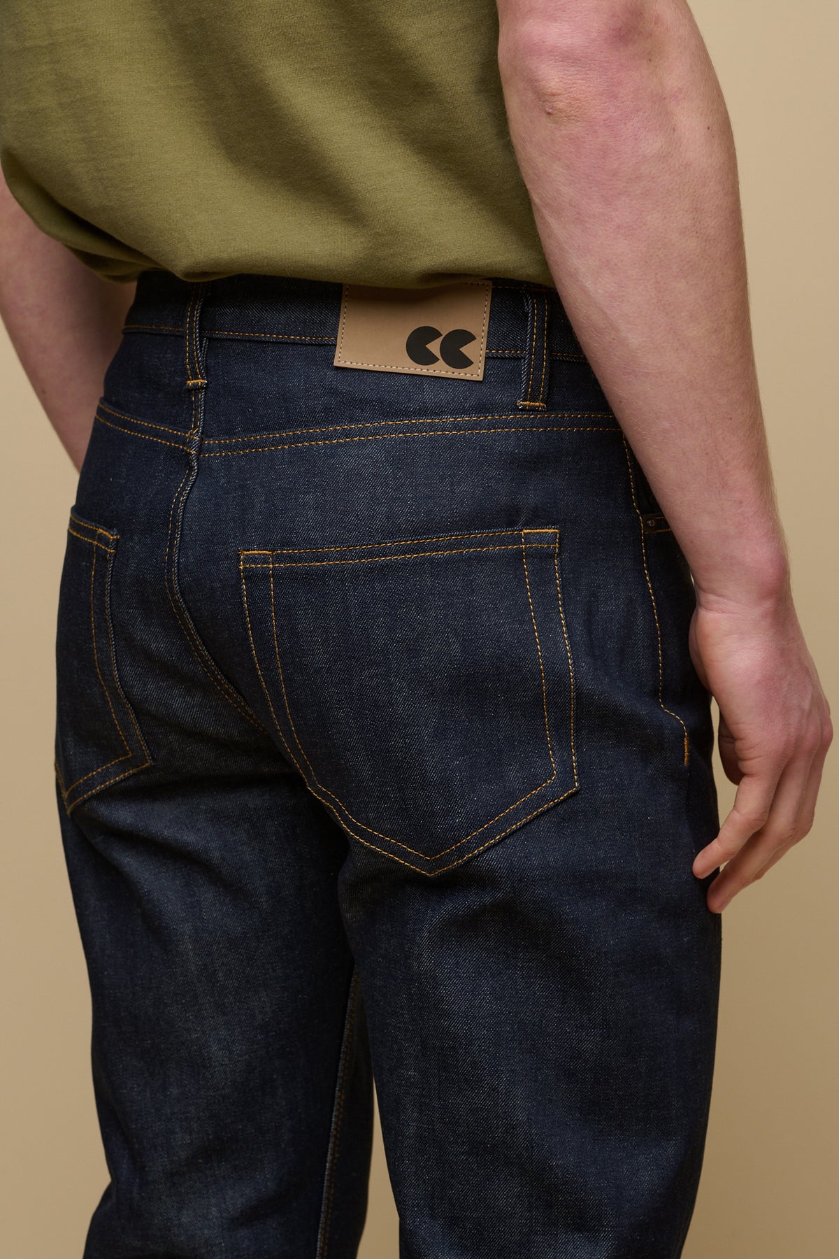 Close up detail shot of the back of men's straight cut selvedge jeans in indigo raw denim with tobacco contrast stitching. Belt loops and CC logo jeans patch on the back of waistband with two back pockets.
