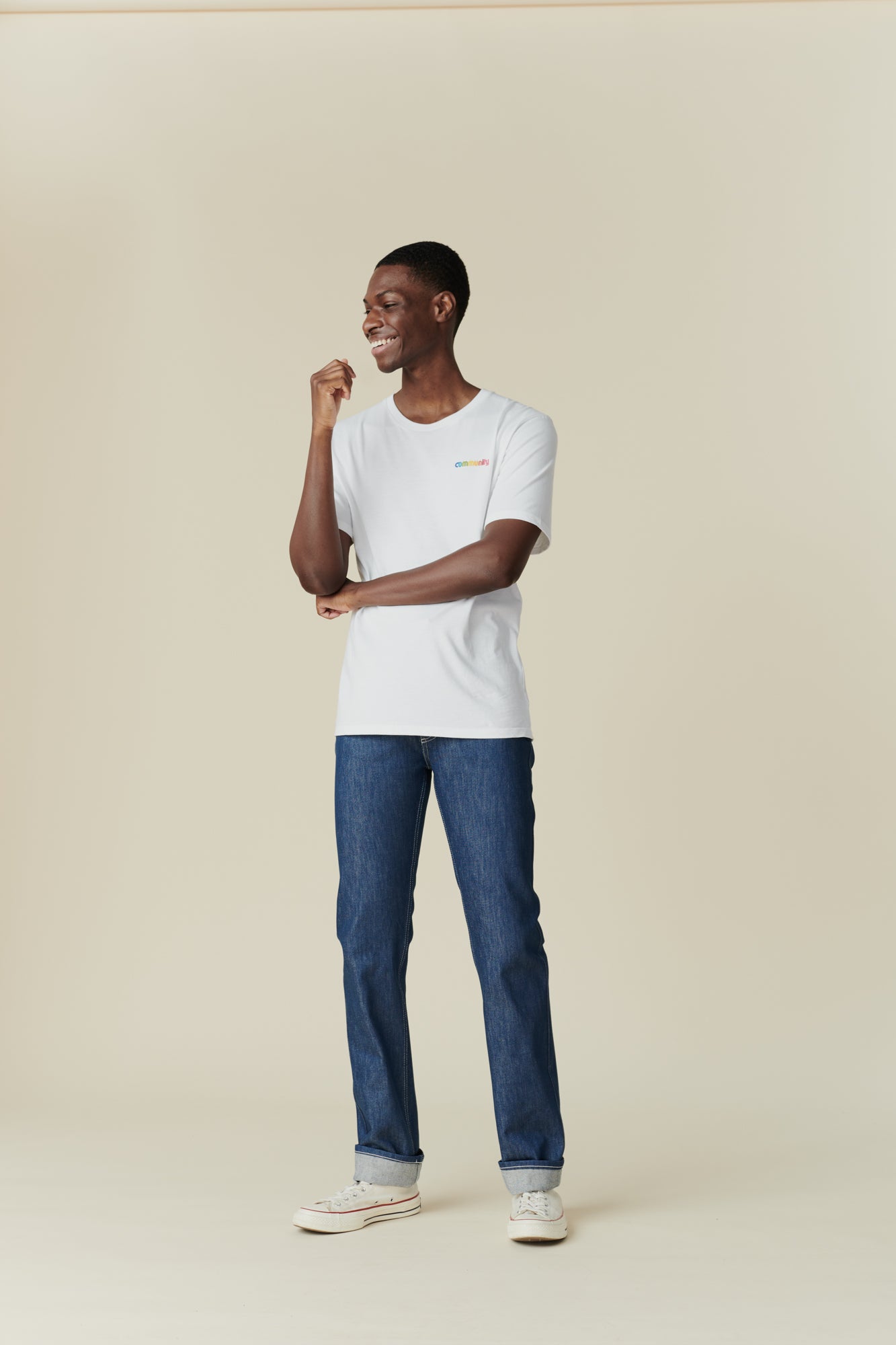 Full body front image of black male wearing straight cut jeans in blue, rolled hem. Paired with white/ multi logo t shirt,