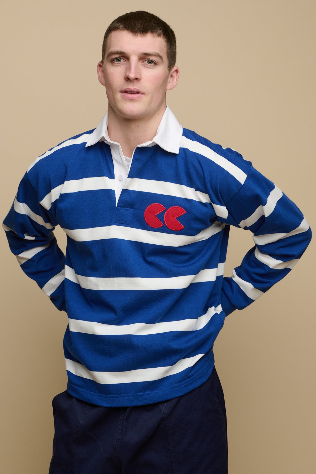 
            Image of brunet male from the thigh up with his hands on the back of his hips wearing blue and white stripe logo rugby shirt with red CC logo patch on chest, worn over crew neck white t shirt