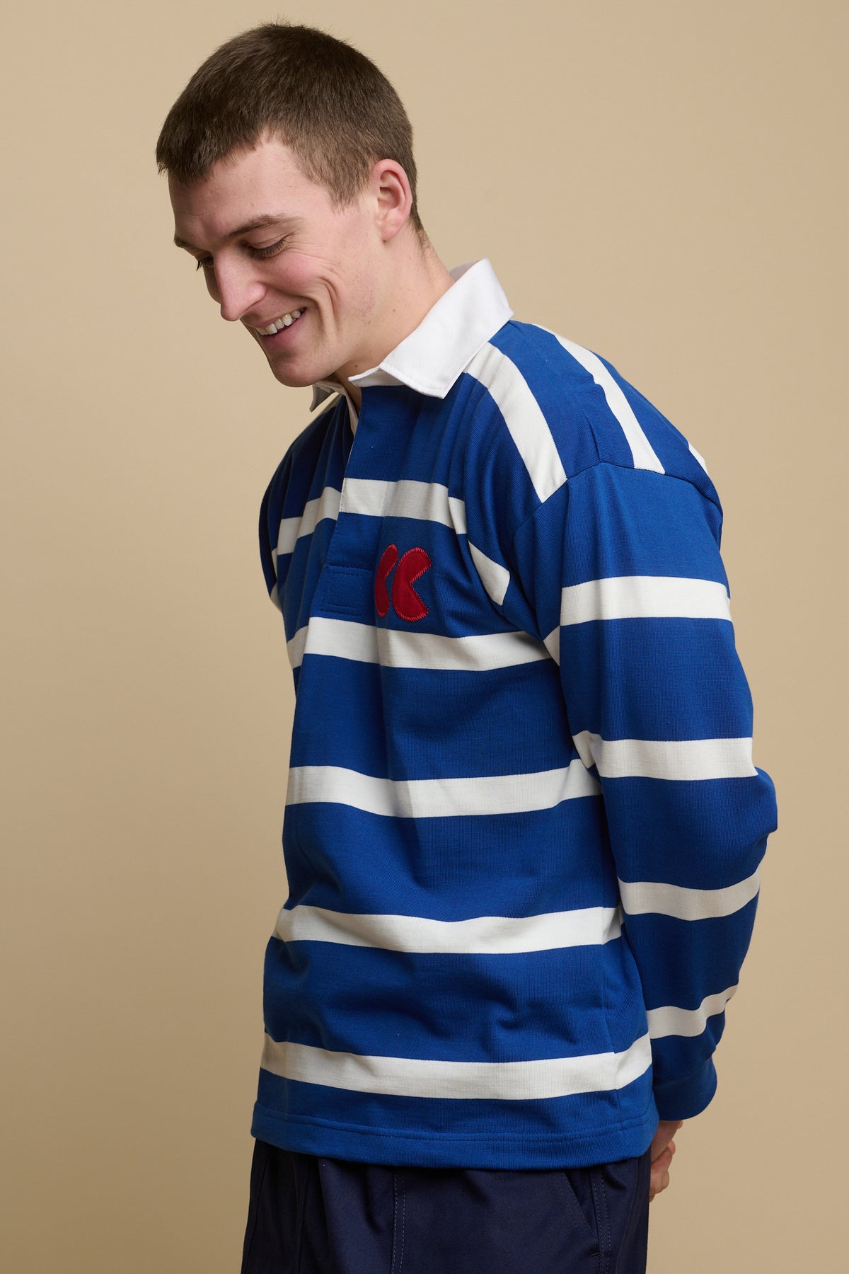 
            Thigh up image of smiley, brunet male wearing stripe logo rugby shirt in blue and white with red CC logo patch on chest