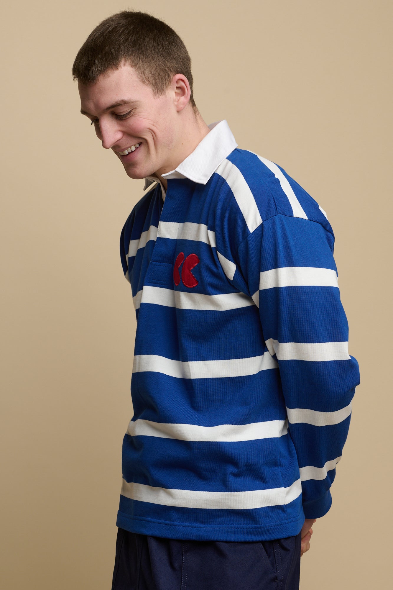 Thigh up image of smiley, brunet male wearing stripe logo rugby shirt in blue and white with red CC logo patch on chest
