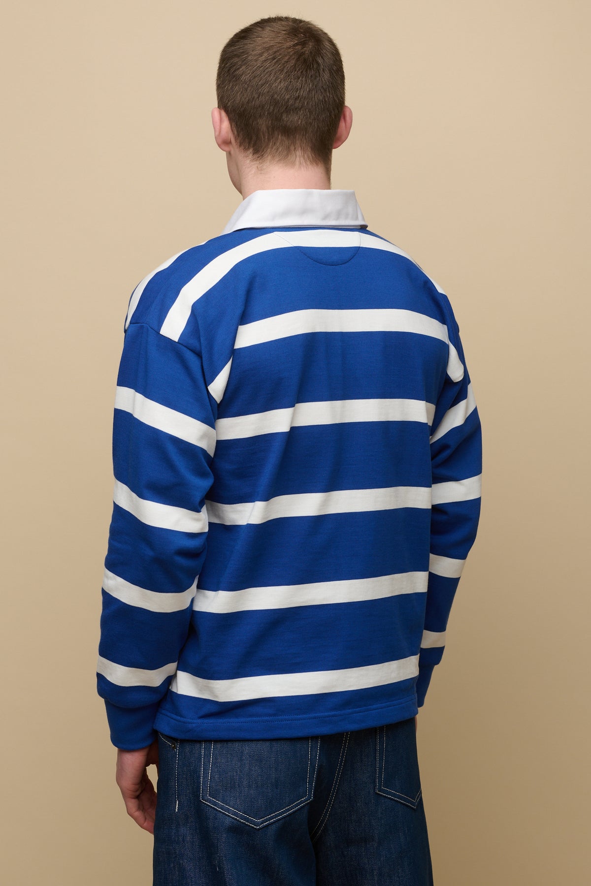 
            The back of male wearing stripe logo rugby shirt in blue white stripes paired with blue jean