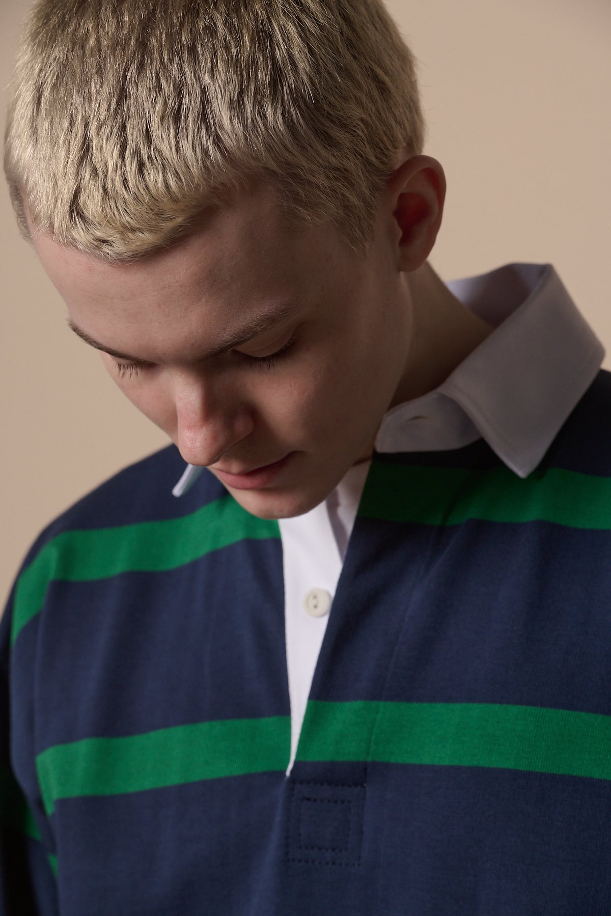 portrait of blonde male looking down wearing navy emerald rugby shirt with white collar.