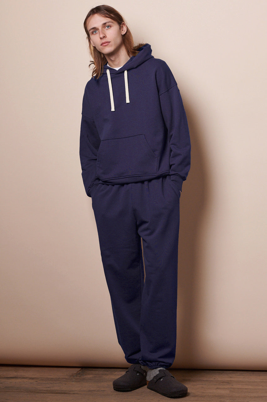 Full body image of male wearing men's sweatpants in navy with both hands in front pockets paired with matching navy hooded sweatshirt
