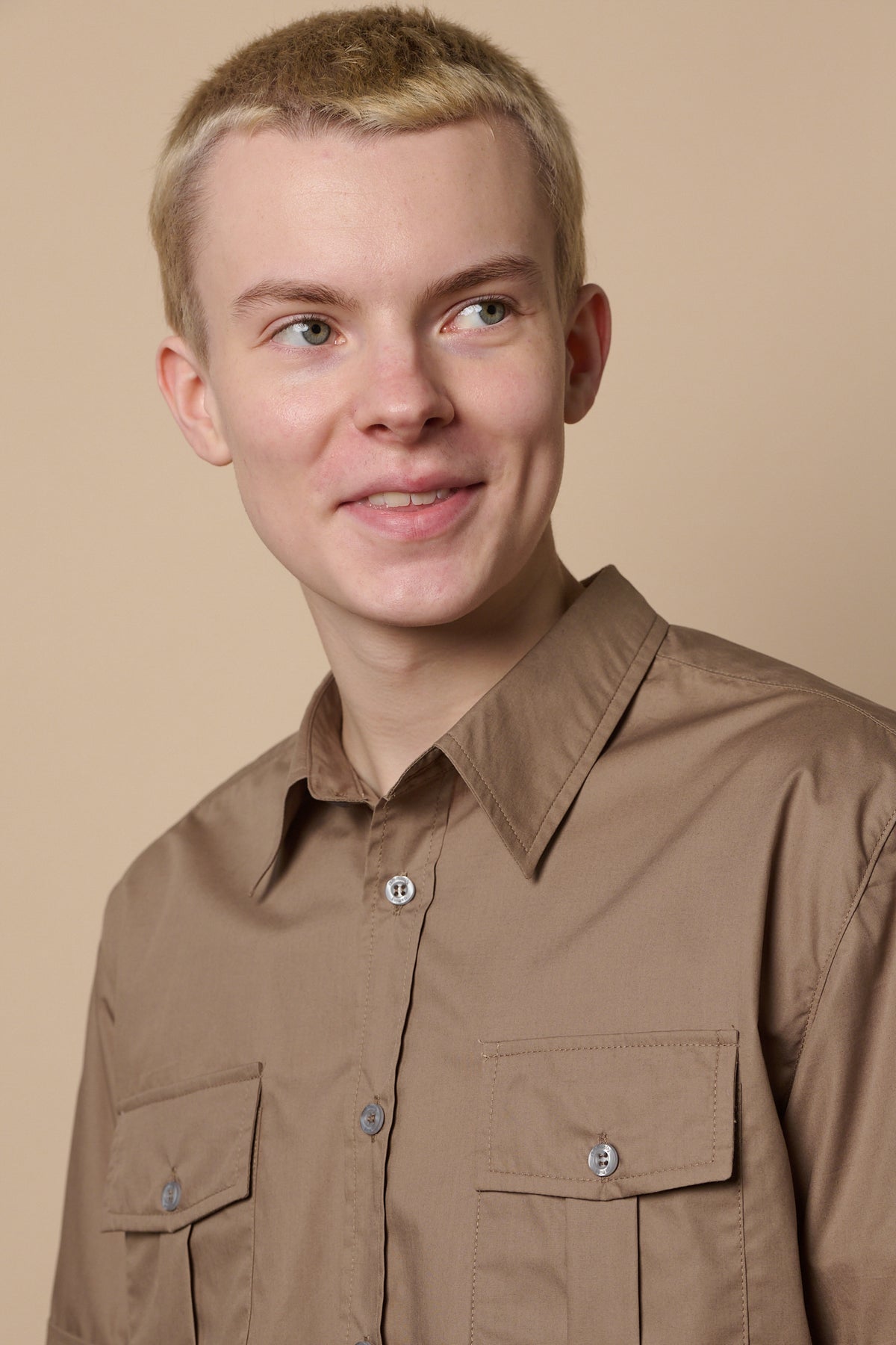 
            Portrait of smirking male with shaved, short blond hair wearing Tom short sleeve military shirt with top button undone.  