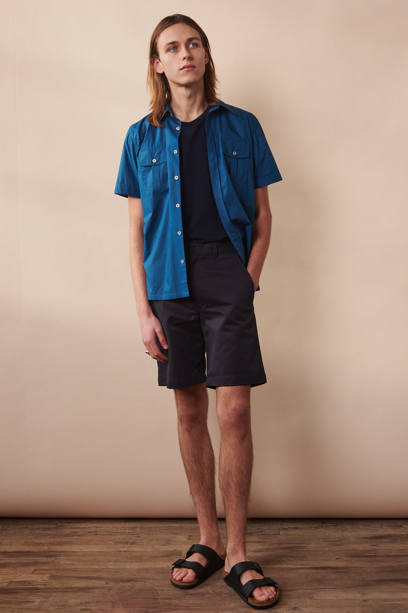 Thigh up image of male sat on stool wearing buttoned Tom short sleeve military shirt in light blue layered over short sleeve t shirt in navy