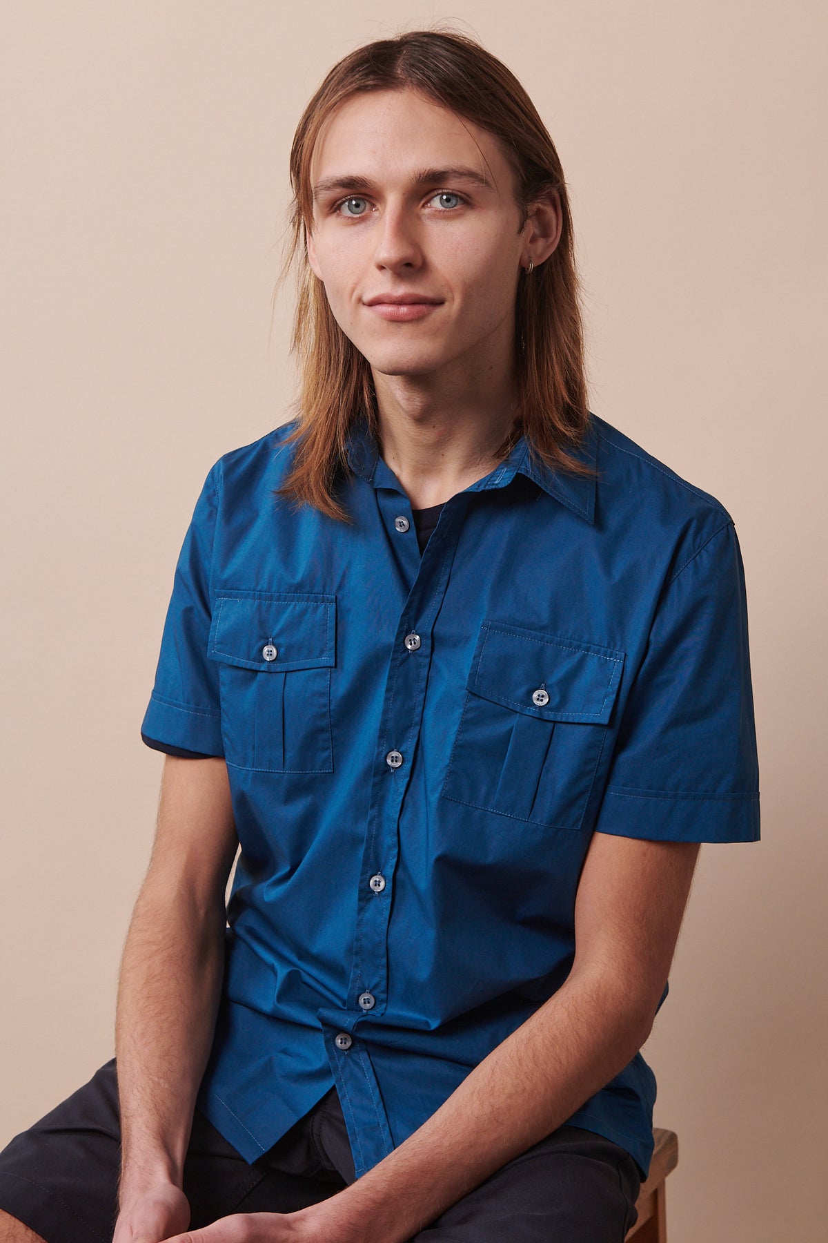 Thigh up image of male sat on stool wearing buttoned Tom short sleeve military shirt in light blue layered over short sleeve t shirt in navy