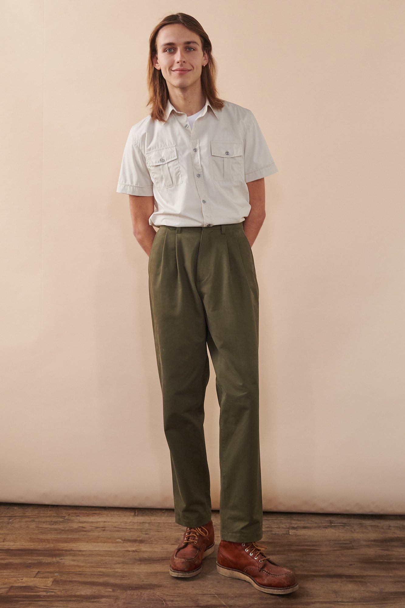 Full body image of male with shoulder length hair holding hands behind back wearing Tom short sleeve military shirt in stone tucked into pleated chino in olive. Top button of shirt undone showing white crew neck t shirt underneath.