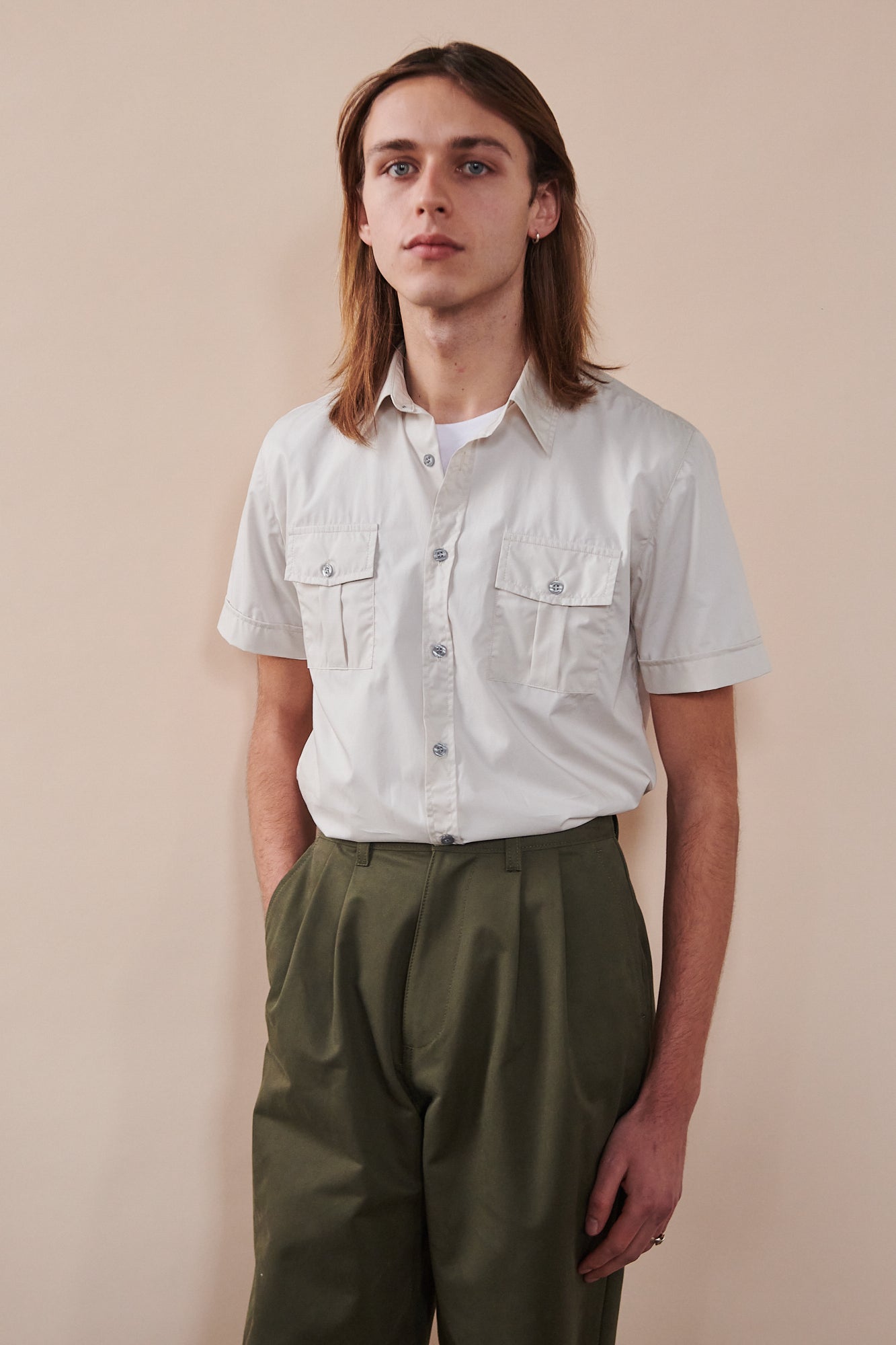 Knee up image of male with shoulder length hair wearing Tom short sleeve military shirt in stone tucked into pleated chino in olive. Top button of shirt undone showing white crew neck t shirt underneath.