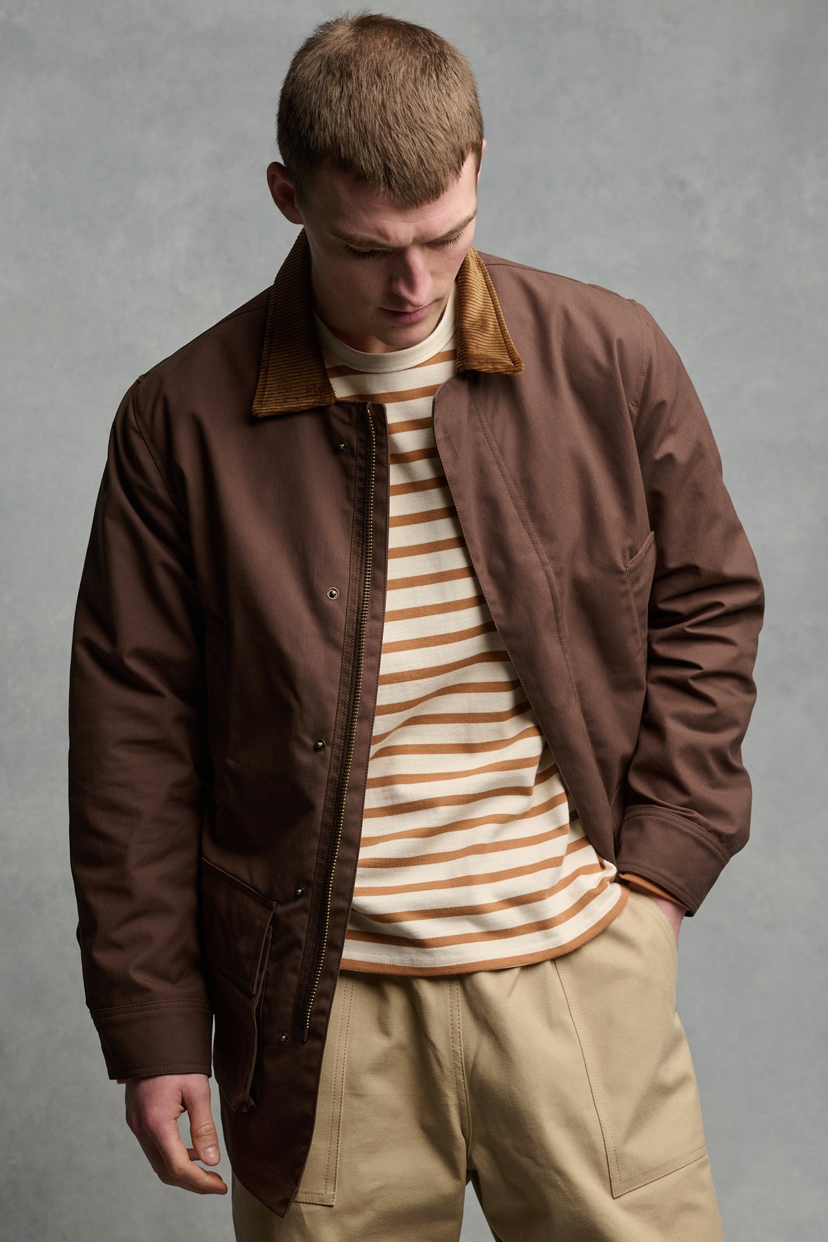 Thigh up image of male wearing men's barn jacket in brown unzipped over Breton in ecru cinnamon paired with cameraman pant in putty