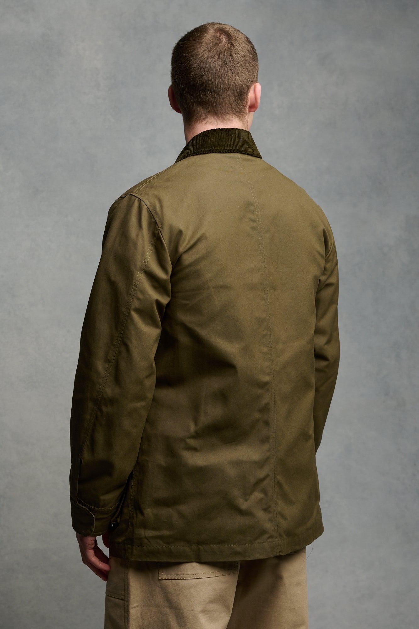 Thigh up image of brunet male wearing unzipped barn jacket in olive worn over sweatshirt in charcoal paired with cameraman pant in putty