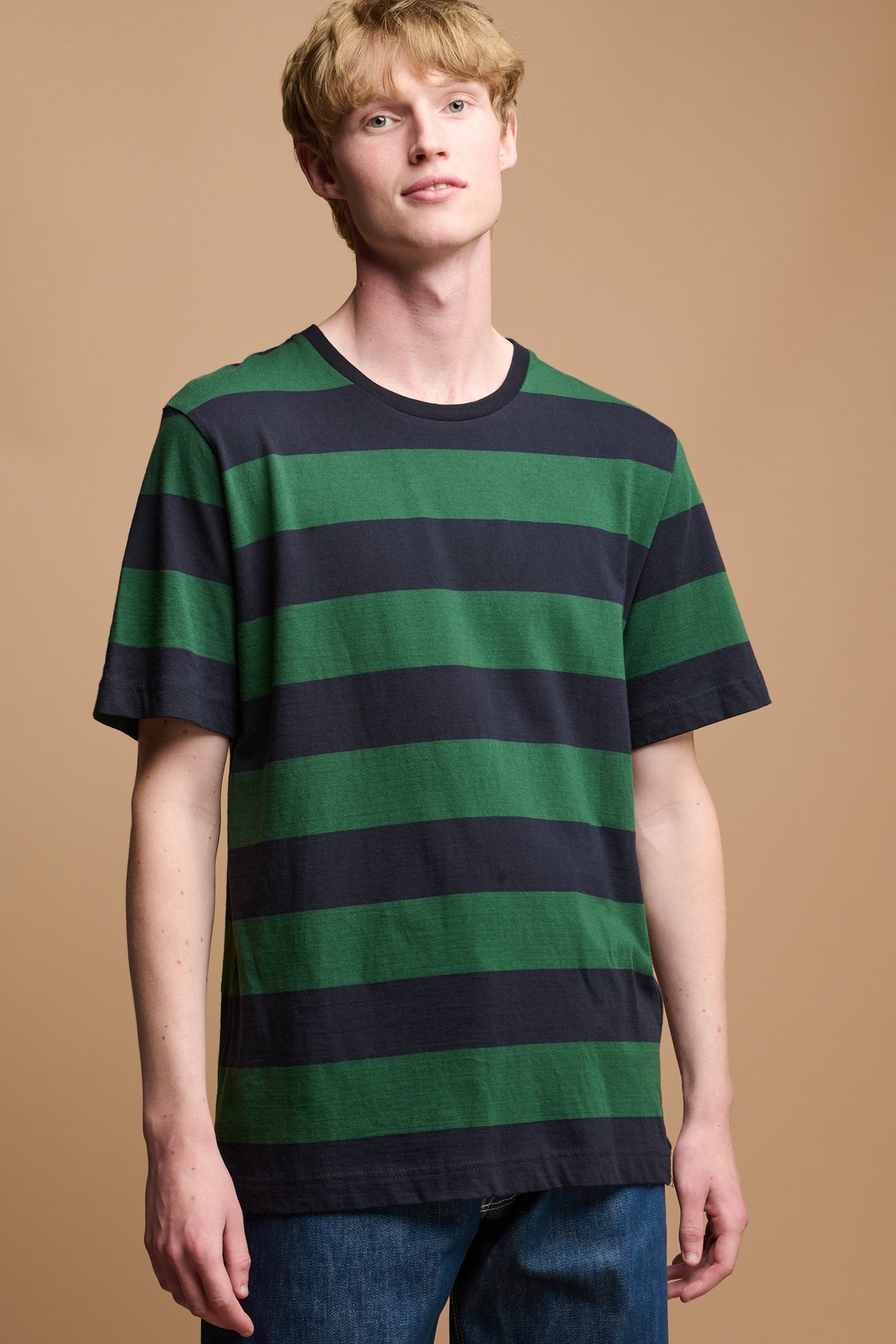 Thigh up image of white male with dirty blonde hair wearing wide stripe t shirt in green and navy stripes