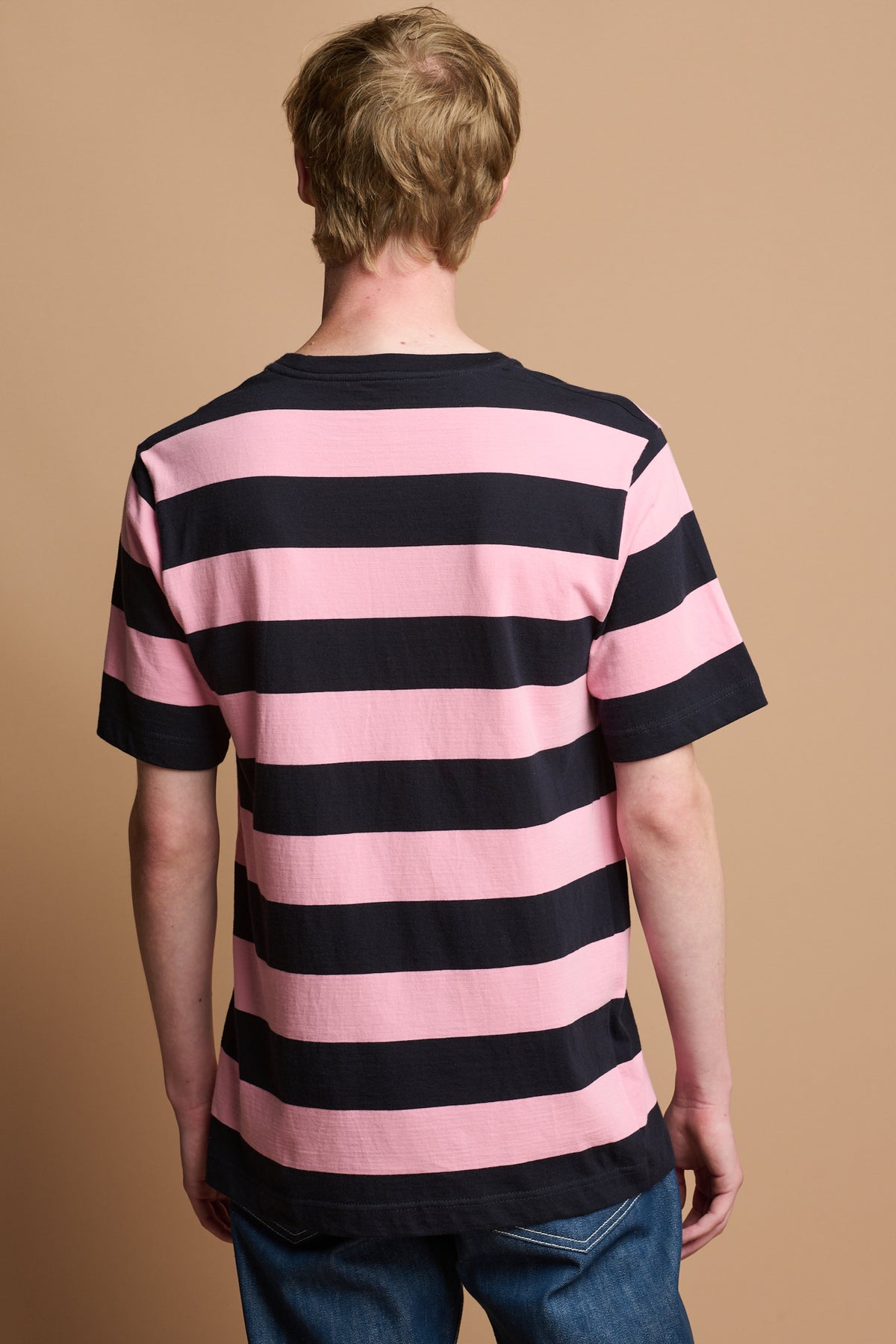 
            the back of male wearing community clothing wide stripe t shirt in pink and navy paired with blue arran button fly jeans