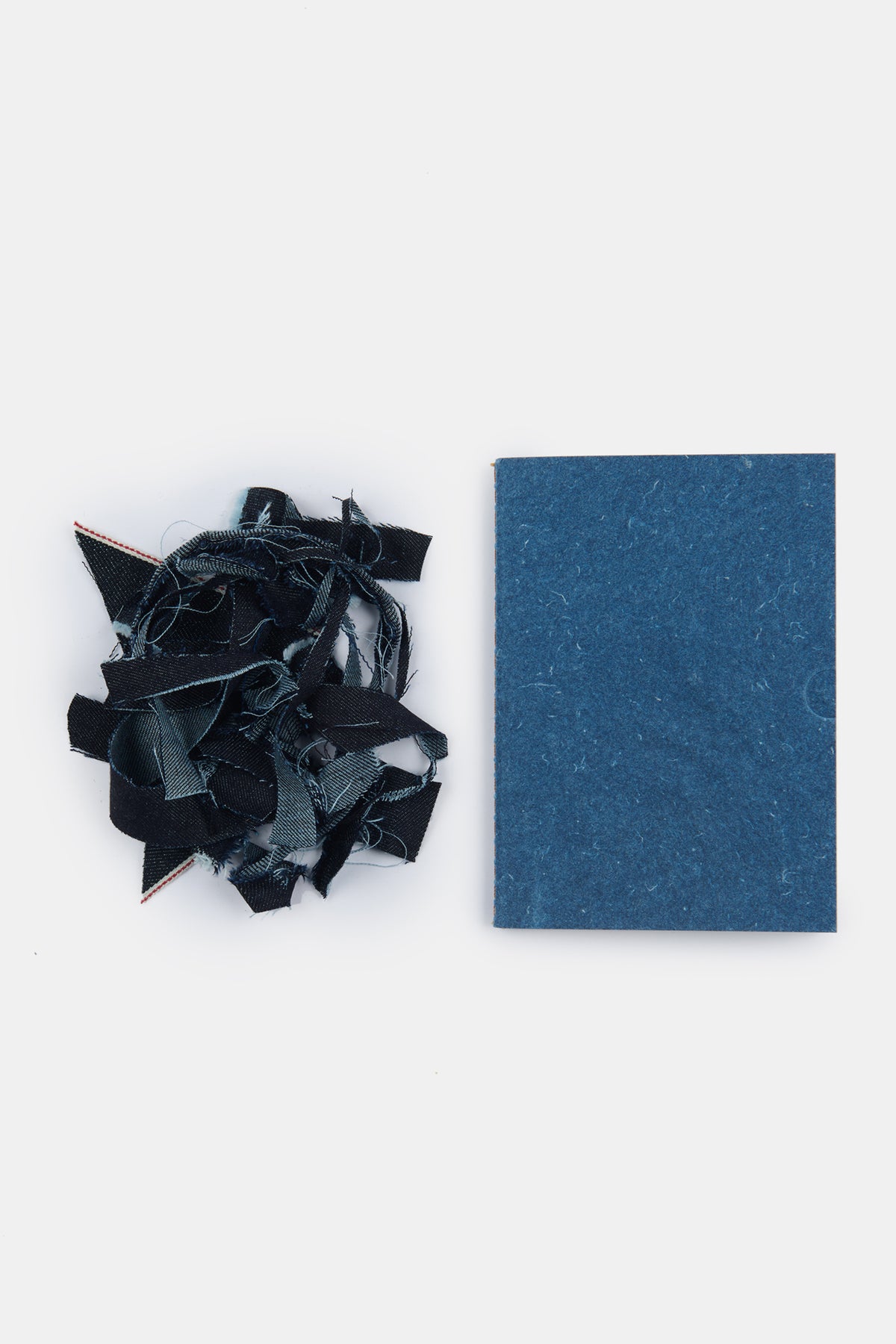 
            The front of Community Clothing x Mark + Fold A6 notebook in indigo lay next to a small pile of indigo denim scraps that the notebook cover is made from