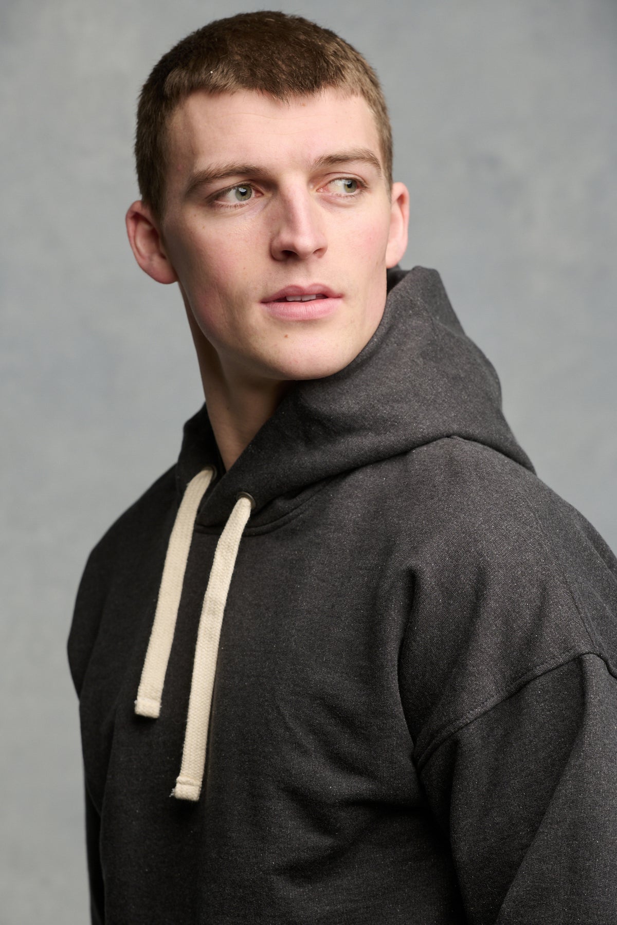 Portrait of the side of male wearing heritage hooded sweatshirt in charcoal with ecru drawcord through the hood
