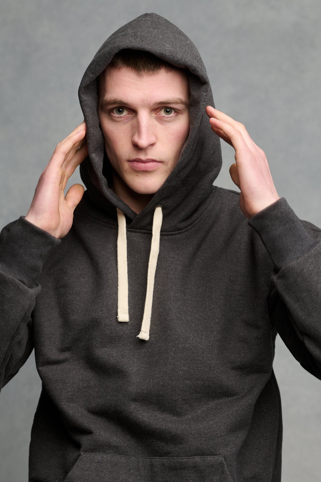 Portrait of male wearing heritage hooded sweatshirt in charcoal with the hood up and hands up towards head. Ecru drawcord through the hood