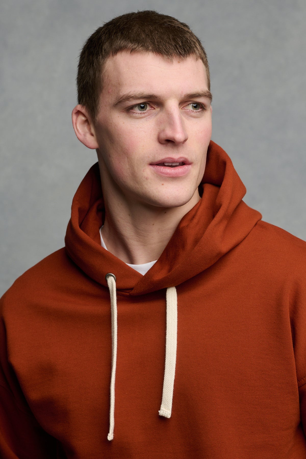Portrait of brunet male wearing men's hooded sweatshirt in cinnamon, worn over crew neck t shirt in white. Ecru drawcord through the hood