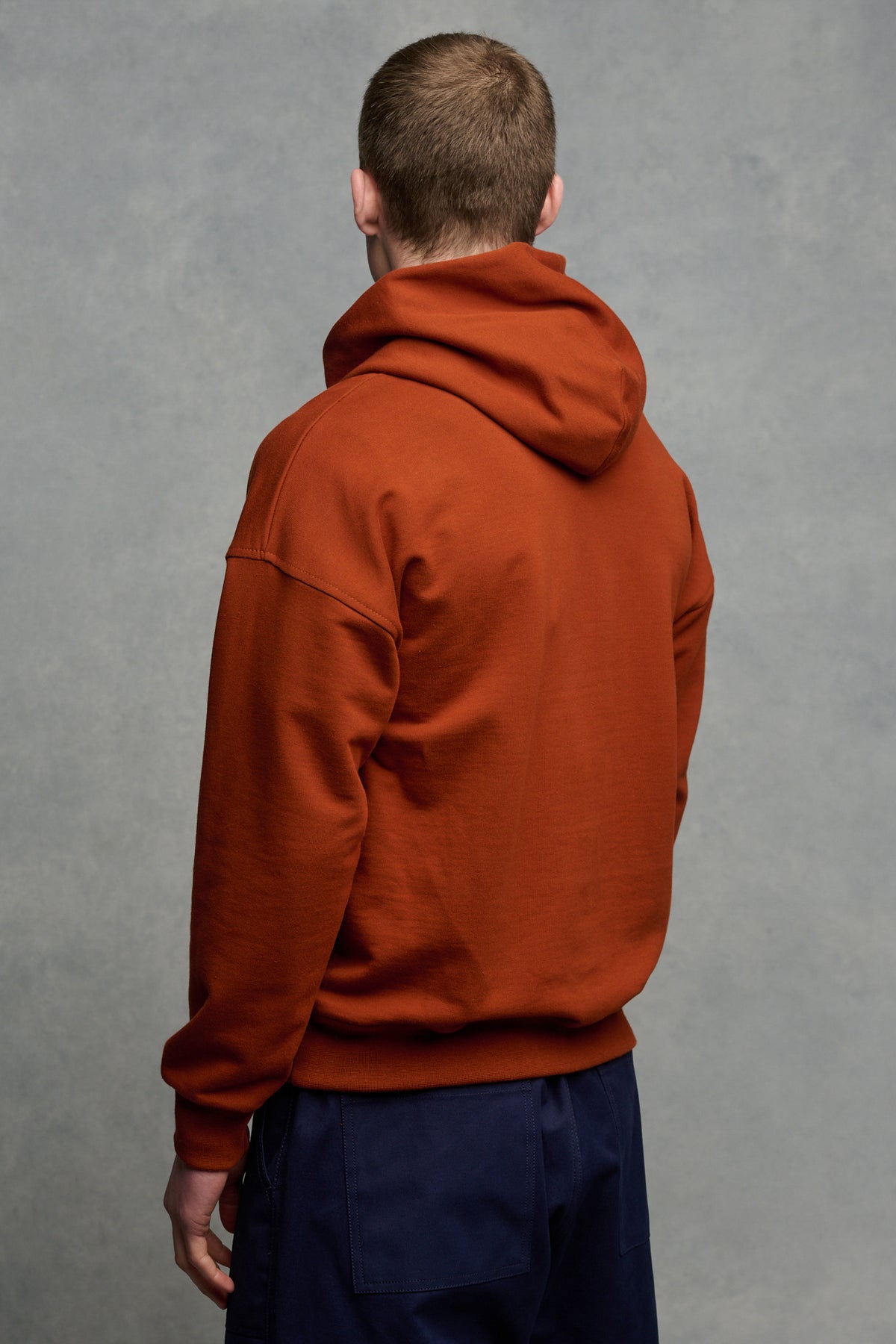 
            The back of brunet male wearing hooded sweatshirt  in cinnamon paired with navy cameraman pant